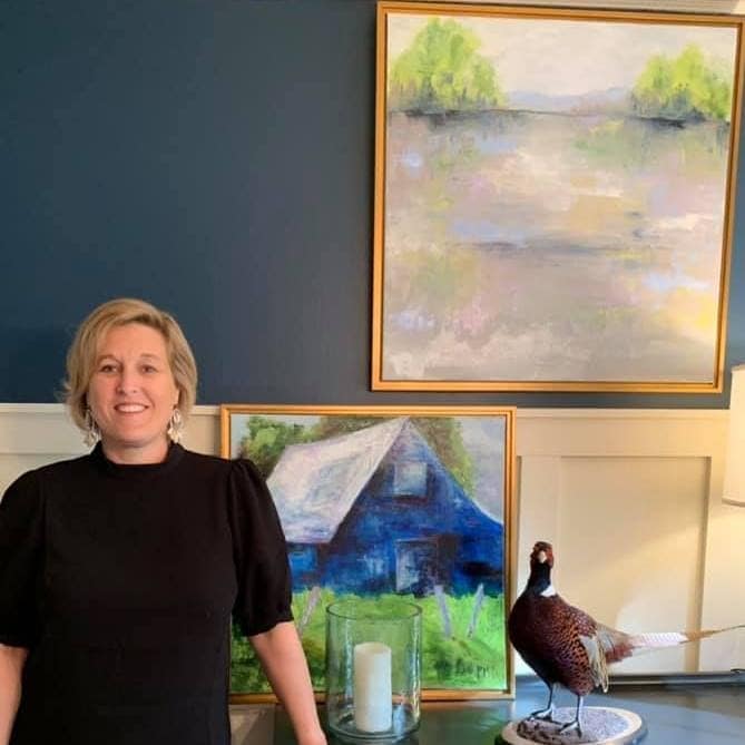 picture of Patti Doyle artist in black shirt woman with blonde hair. standing in front of two framed landscape paintings