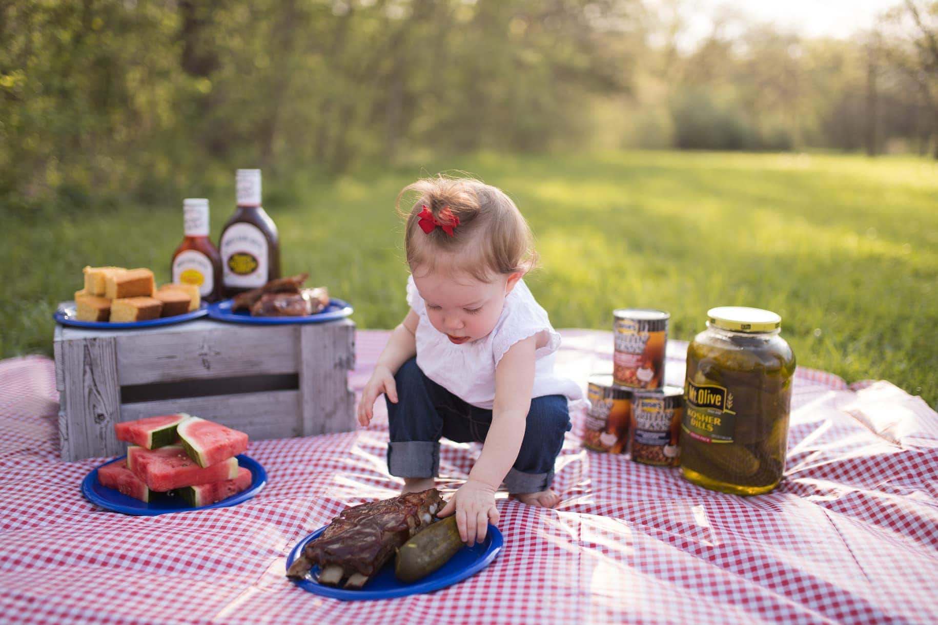 baby with BBQ picnic on a picnic blanket in the grass. baby reaches for pickle on plate