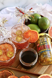 Two cocktail glasses with glitter confetti decoration coming out of the top. Cocktail liquid is orange and there are 2 jalapenos in each glass with grapefruit slice garnish. bowl of limes and jar of Sweet Heat Jalapenos shown