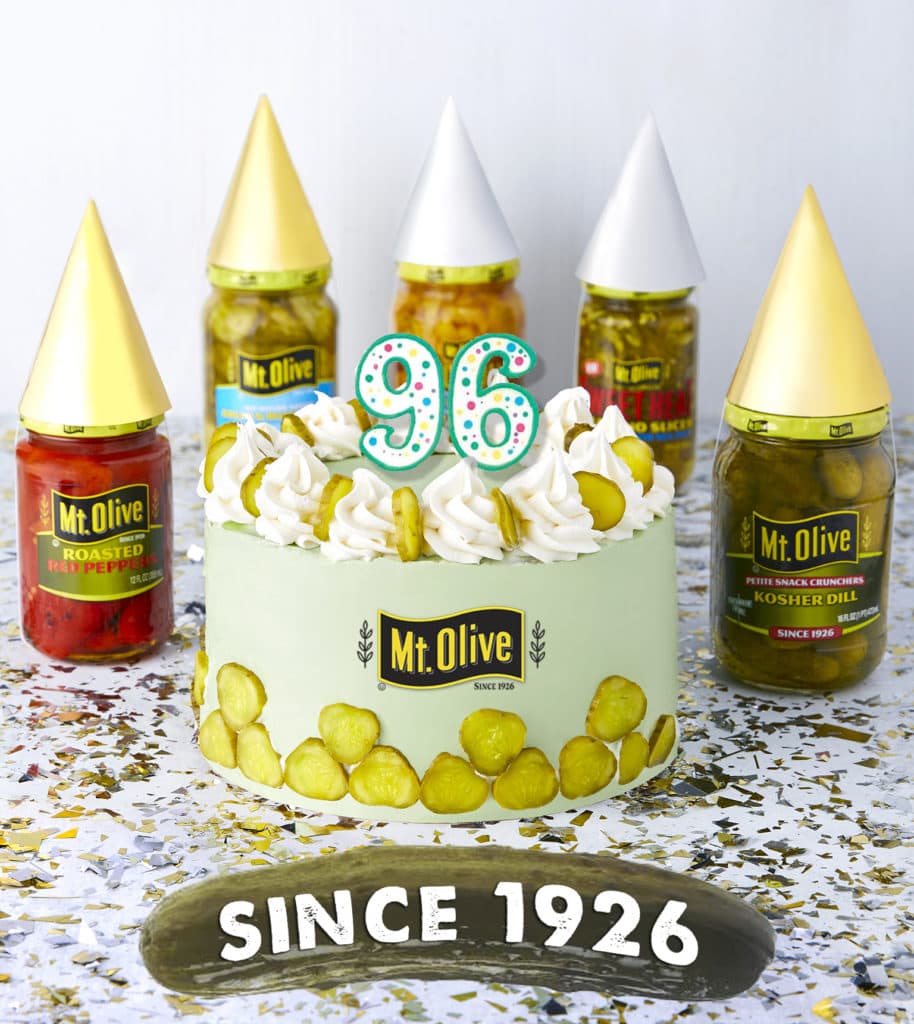 Birthday cake with candles that say 96. green cake with pickles on the bottom. Jars of Mt. Olive products surrounding. pickle banner says Since 1926