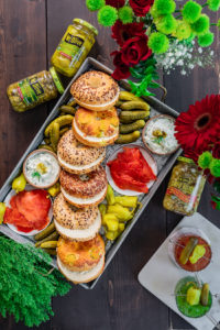 Wood table with gray tray. On tray is an assortment of bagels with smoked salmon lox. pickles shown on tray. Small dishes of cream cheese. Surrounded by jars of pickles and red and green flowers.