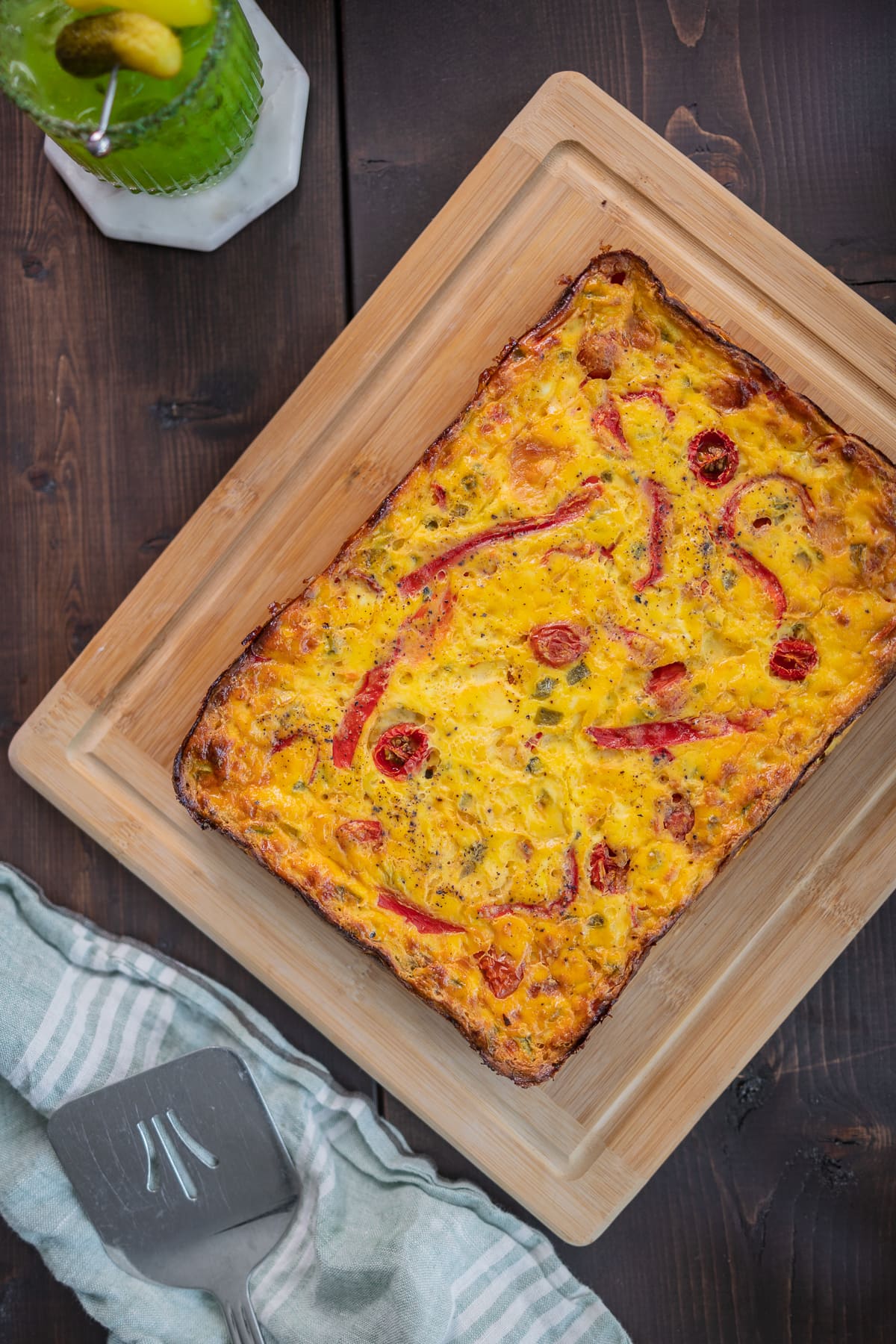 dark wood table with light wood board. on board is a veggie and egg frittata with tomatoes and red peppers
