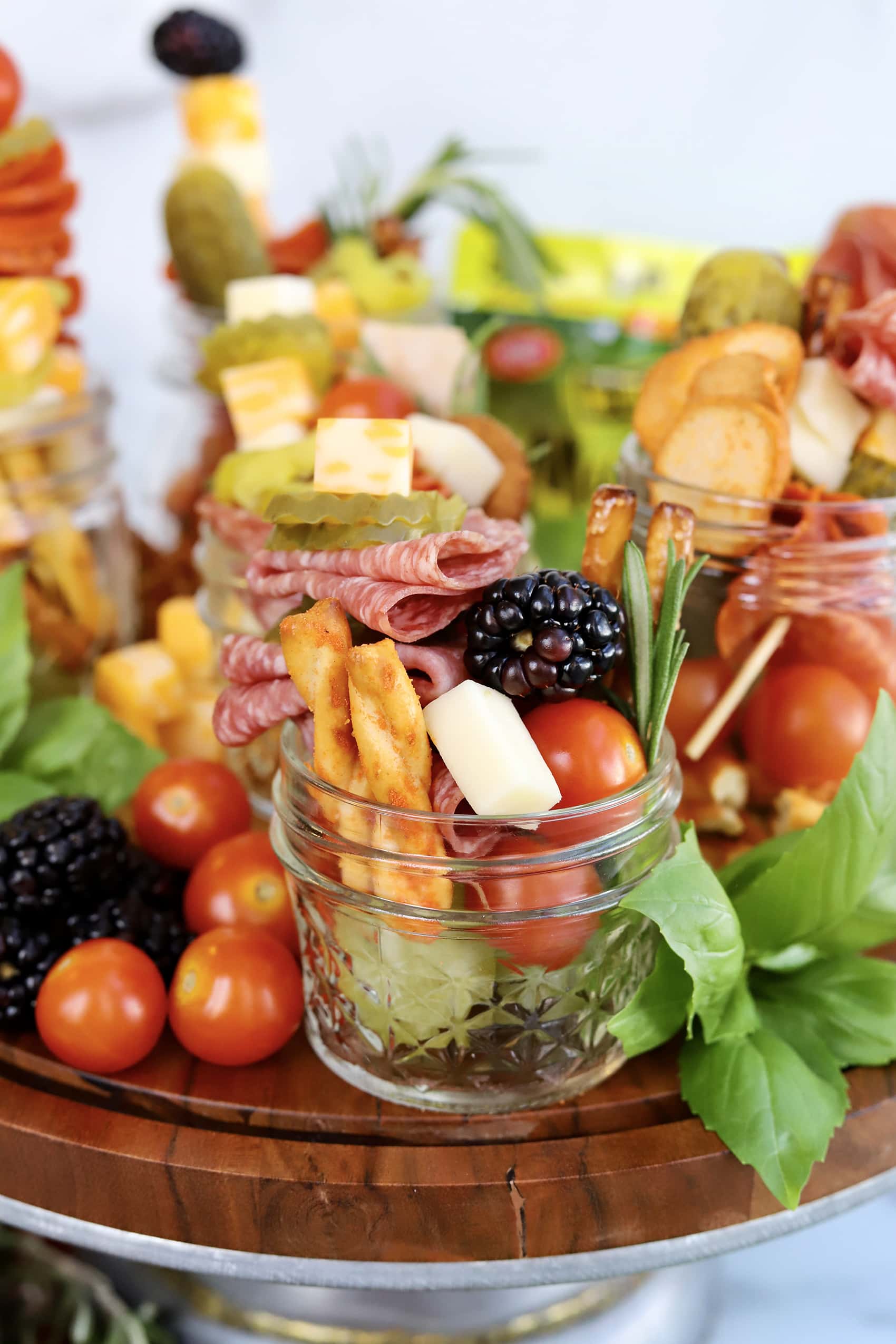 small glass jar filled with pretzels, berries pickles, cheese and tomatoes shown on wooden display board surrounded by charcuterie
