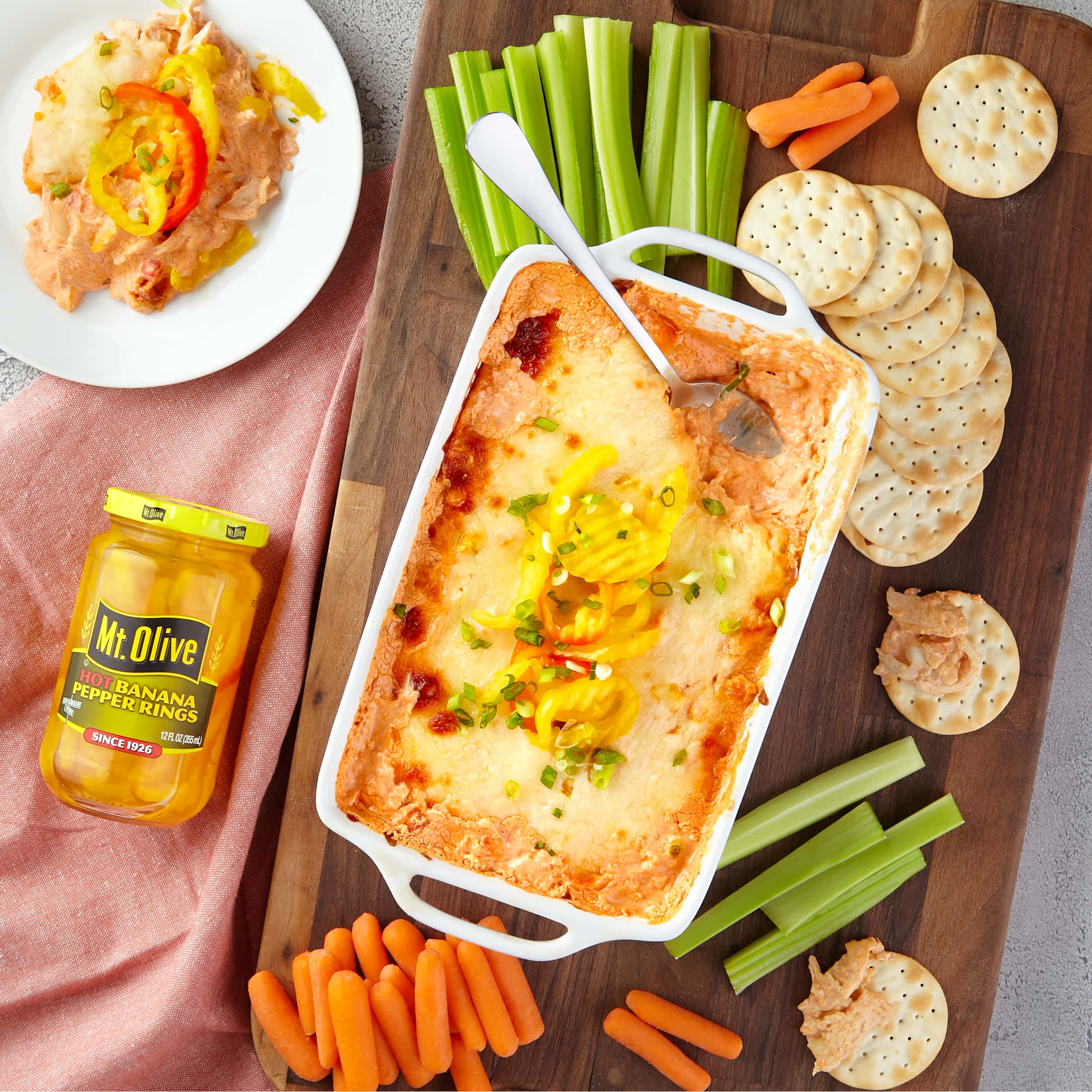 wood cutting board with celery and carrots and crackers. White dish on top with buffalo chicken dip topped with cheese and banana peppers. Shown next to it is a jar of Hot Banana Peppers.
