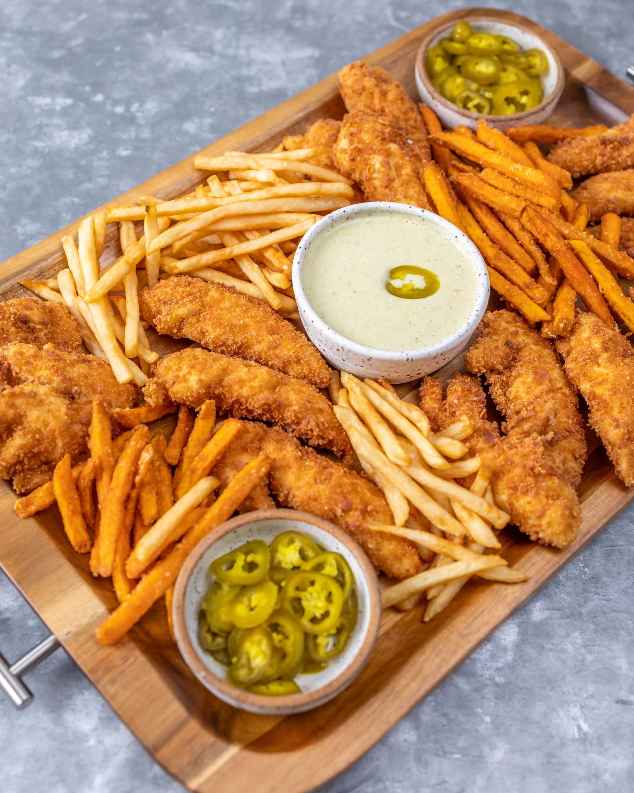 grey countertop. wood tray with dishes of jalapenos and ranch dressing. tray shows french fries and chicken tenders.