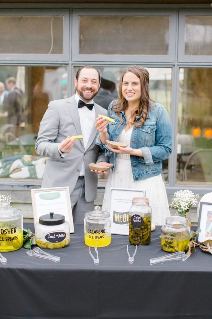 Bride in white dress with jean jacket stands next to groom in grey jacket with black tow tie. Both are holding pickle spears. table filled with jars of pickles in front of them.