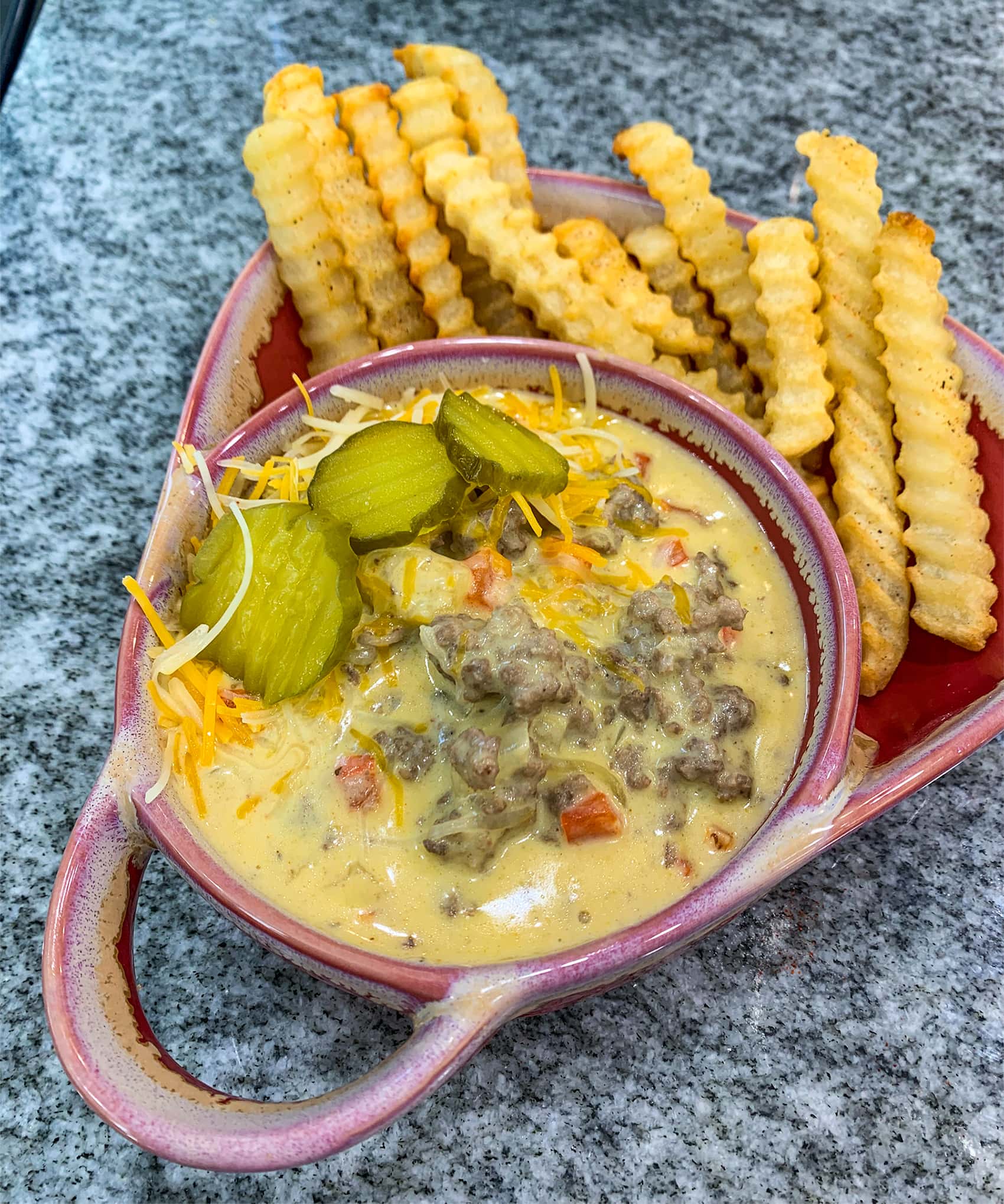 black & white countertop with a plate and bowl. plate has crinkle cut french fries. bowl shows creamy cheesy soup with ground beef and pickles on top.