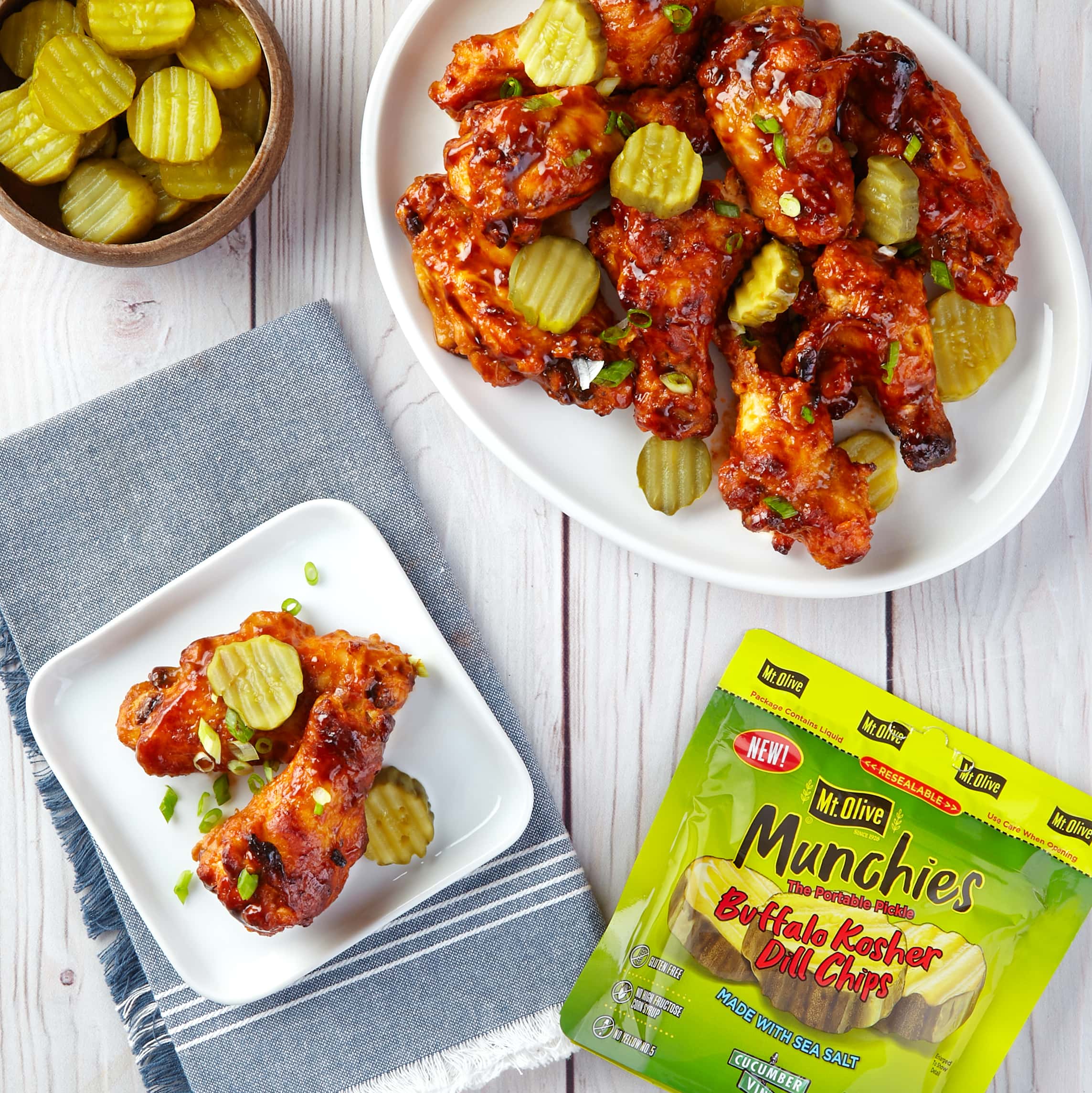 white wood table. shows pack of green Mt. Olive Munchies Pickle Pouch Buffalo Chips and a large white plate filled with chicken wings and a small white plate with 2 chicken wings. Wings are topped with pickles