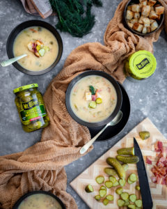 Bowl of pickle soup with croutons on top. Jar of Kosher Baby Dill pickles. Cutting board with pickles and bacon chopped up.