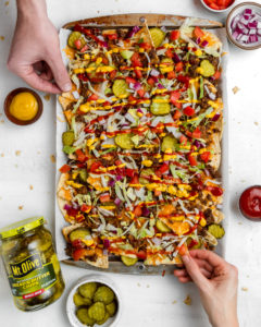 large serving of cheeseburger nachos (chips, cheese, ground beef, ketchup, mustard and pickles) with jar of pickles on the side and dish of pickle chips