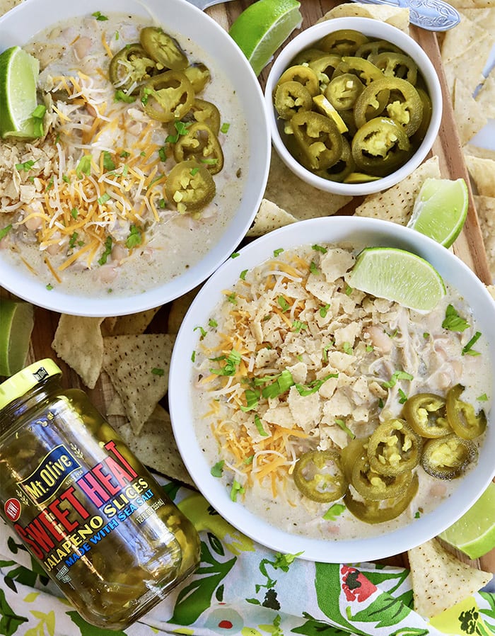 two bowls of white chicken chili with a dish of jalapeños and a jar of Sweet Heat Jalapnos. Chili is topped with jalapeños.