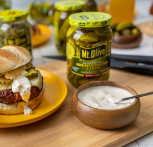 wood cutting board showing jar of pickles and a brown wooden dish of white sauce which is the Dill Mustard Burger Sauce. Yellow plate with a burger next to it. burger topped with pickles and sauce