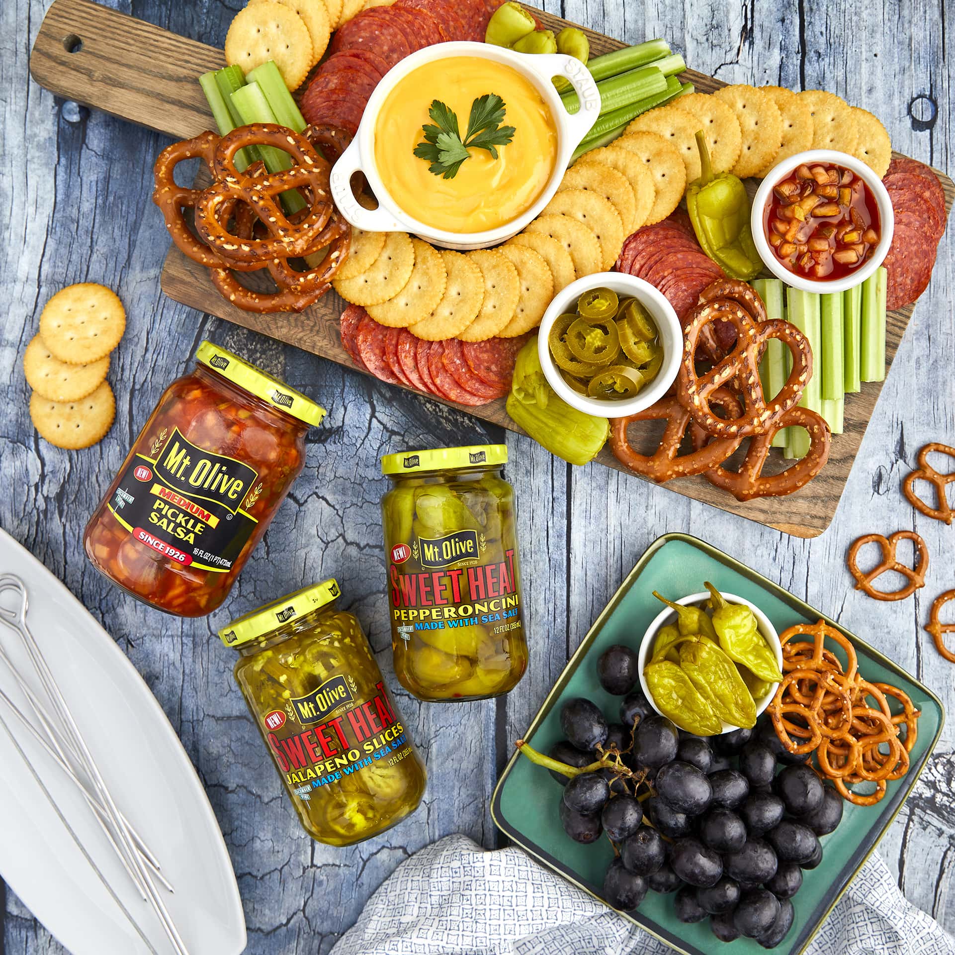 jars of Pickle Salsa, SweetHeat Pepperoncini and SweetHeat Jalapenos. Charcuterie board made of wood shows crackers, pretzels, cheese dip and dishes of pickle salsa and peppers.