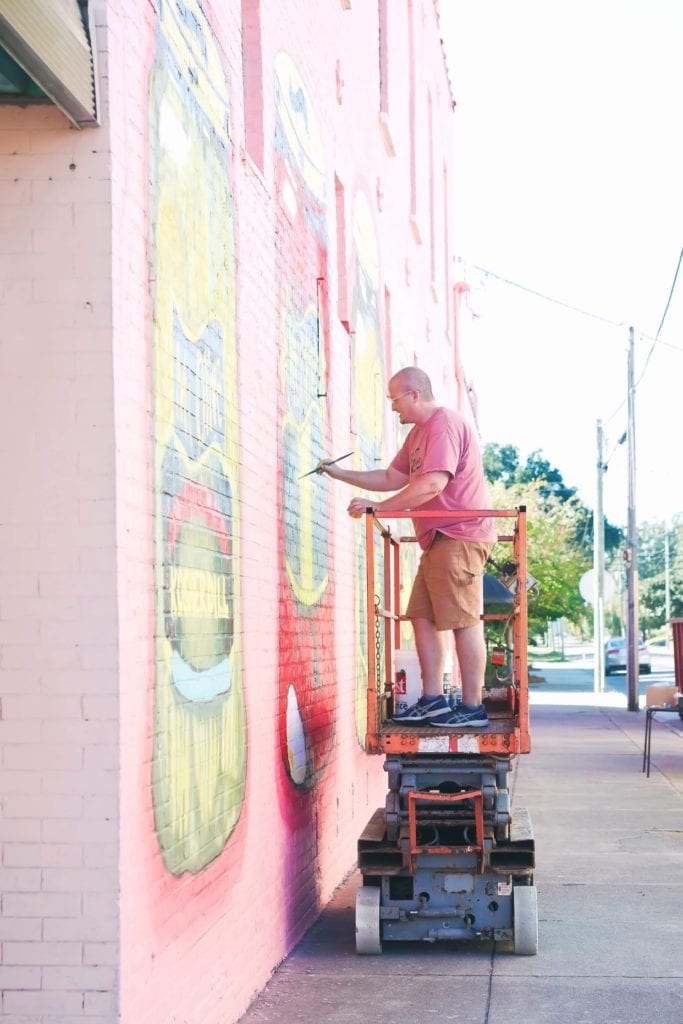 Artist creating colorful mural on brick wall for Mt Olive Pickles.