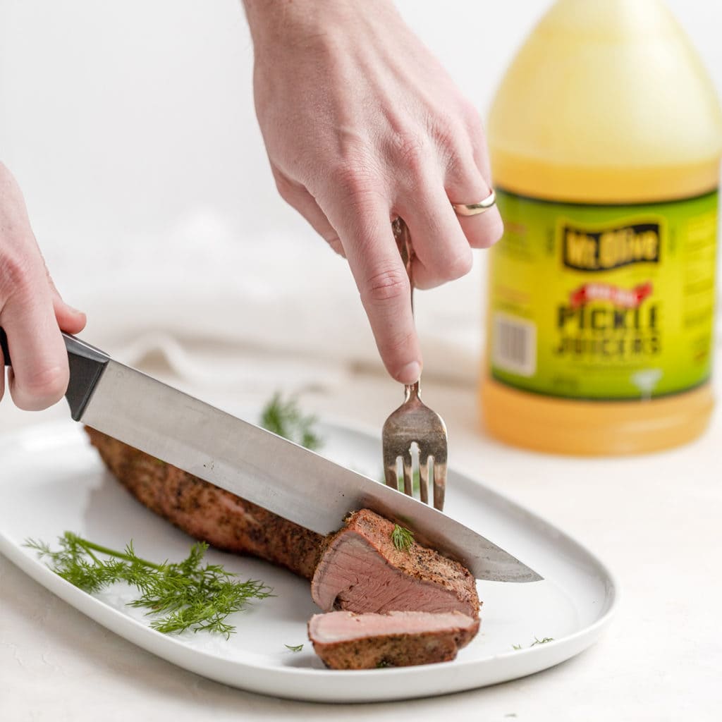 pork tenderloin on white plate. person cutting into the tenderloin. 64 oz jug of Pickle Juicers in background