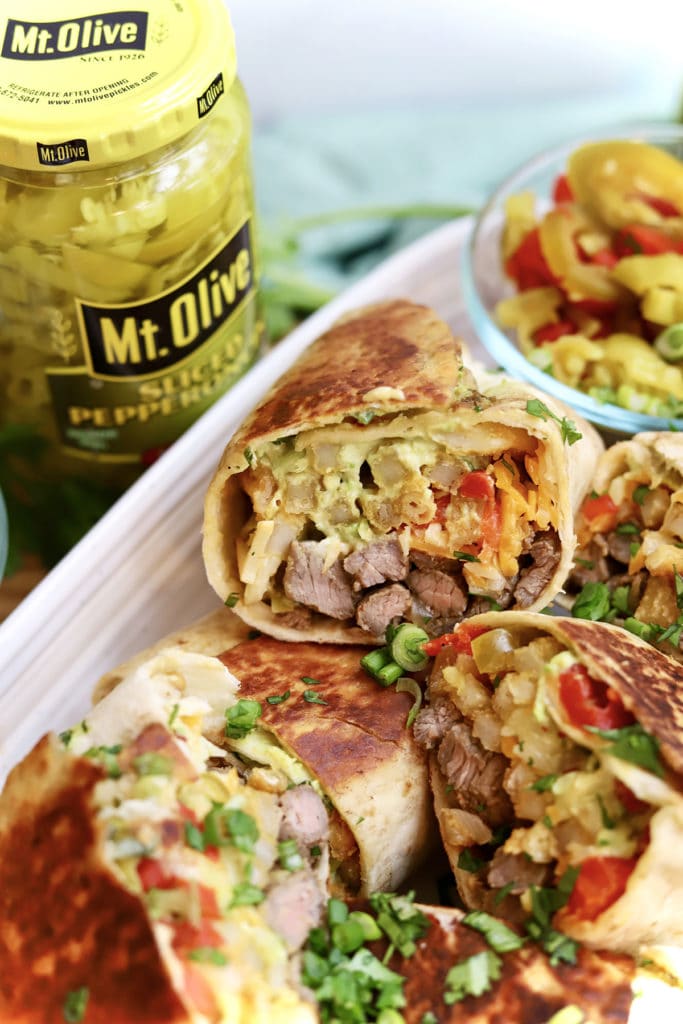 Jar of sliced pepperoncini on the left. dish with a close up of a burrito cut in half. burrito contains steak and peppers and avocado cream sauce. topped with green onions.
