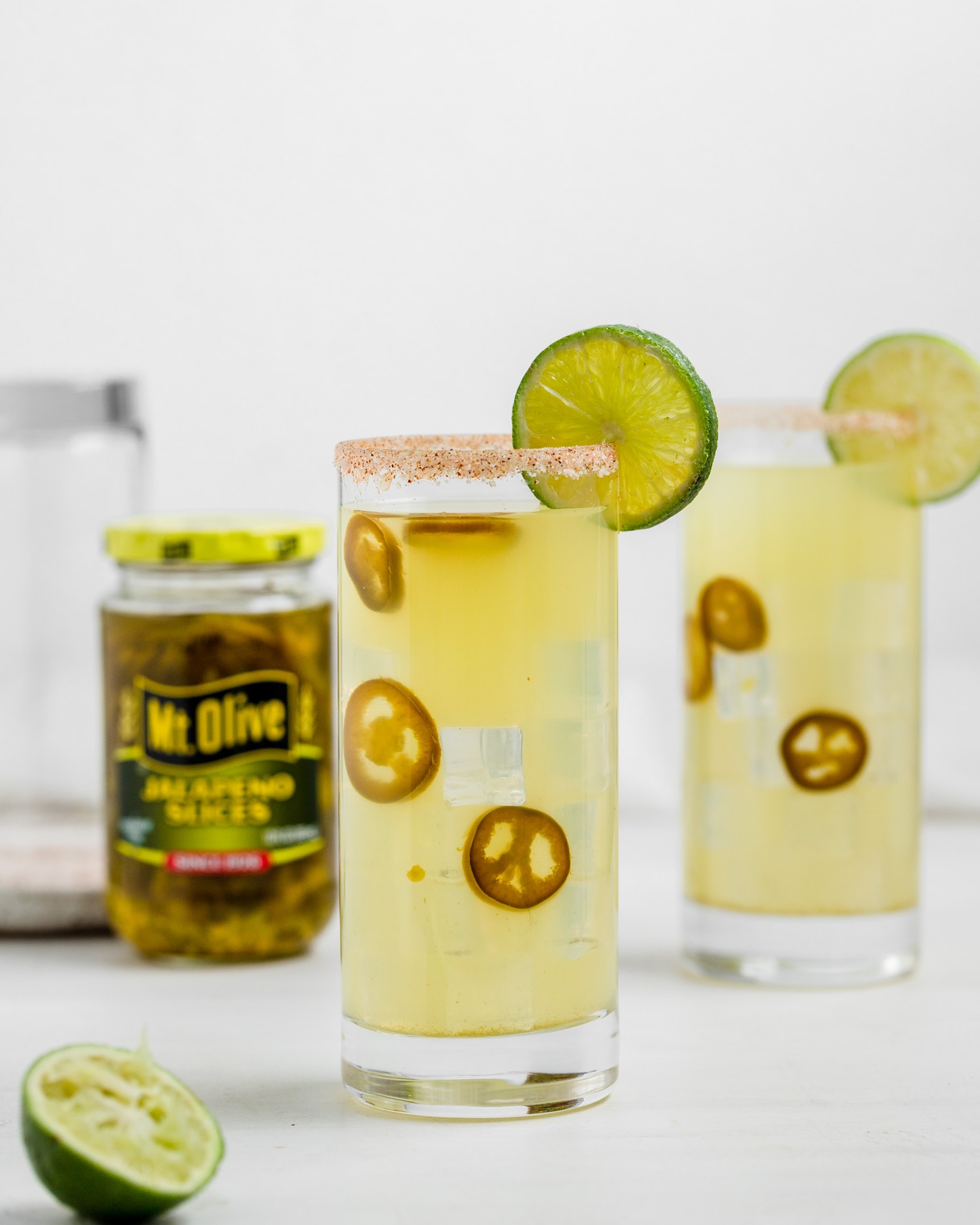two glasses with margarita liquid (yellow) and jalapeno slices in the drink- garnished with lime slices. jar of Mt. Olive Jalapeno Slices in background.