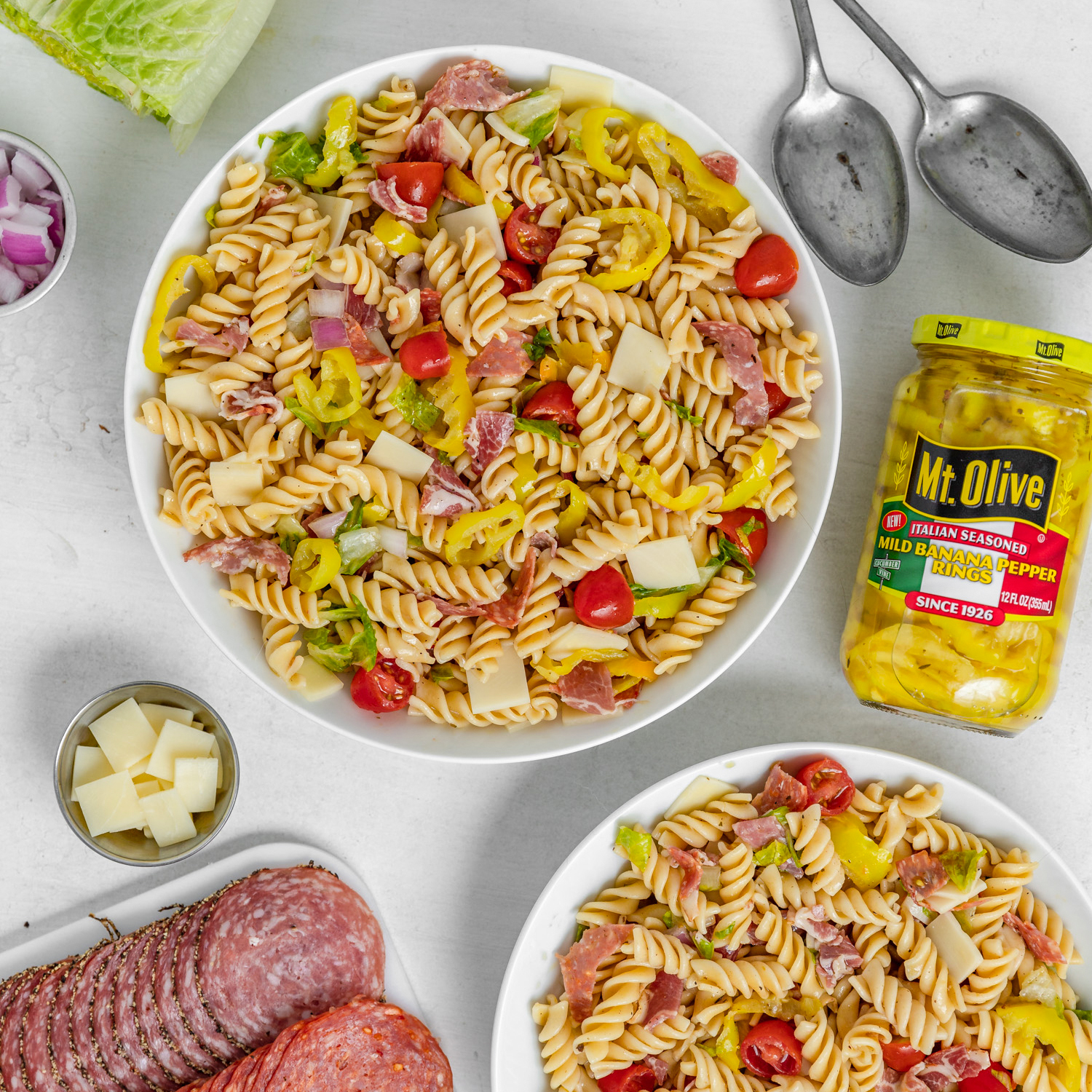 two white bowls filled with pasta salad, tomatoes, banana peppers, cheese and salami. jar of Italian Seasoned Banana Peppers shown.