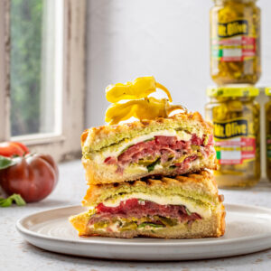 Large stacked panini sandwich with meat, cheese, peppers and jars of peppers in background. tomato on the side