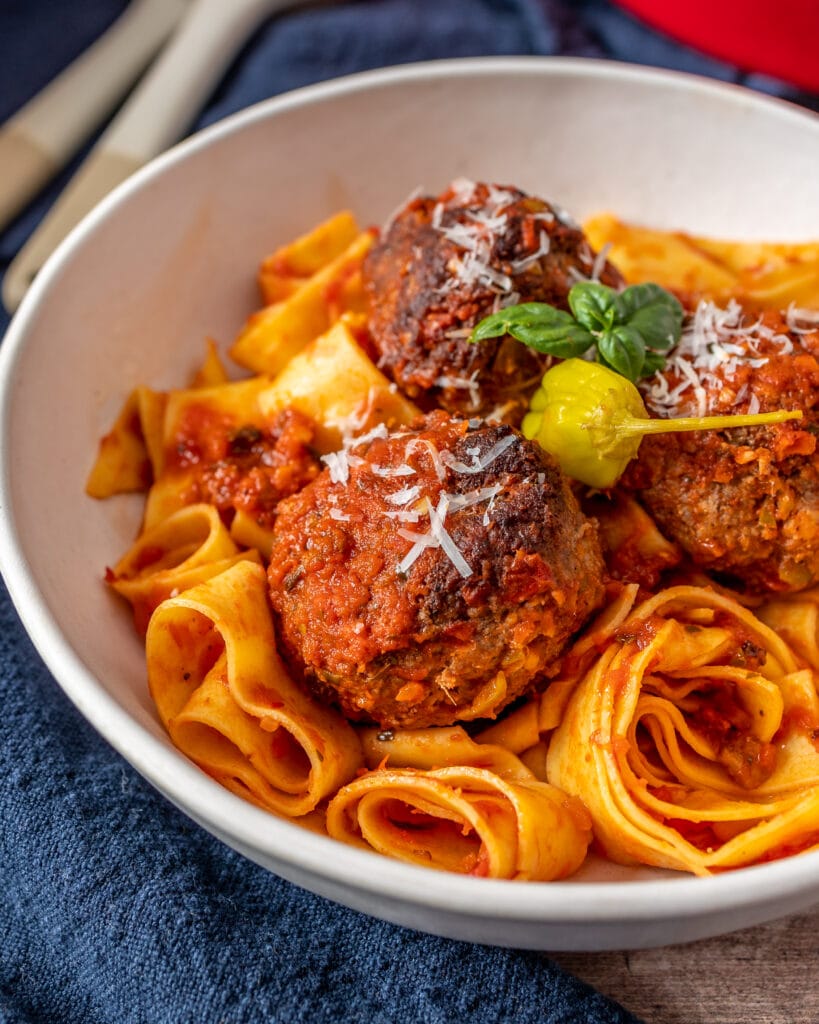 white bowl of meatballs with pasta and pepperoncini and basil as garnish