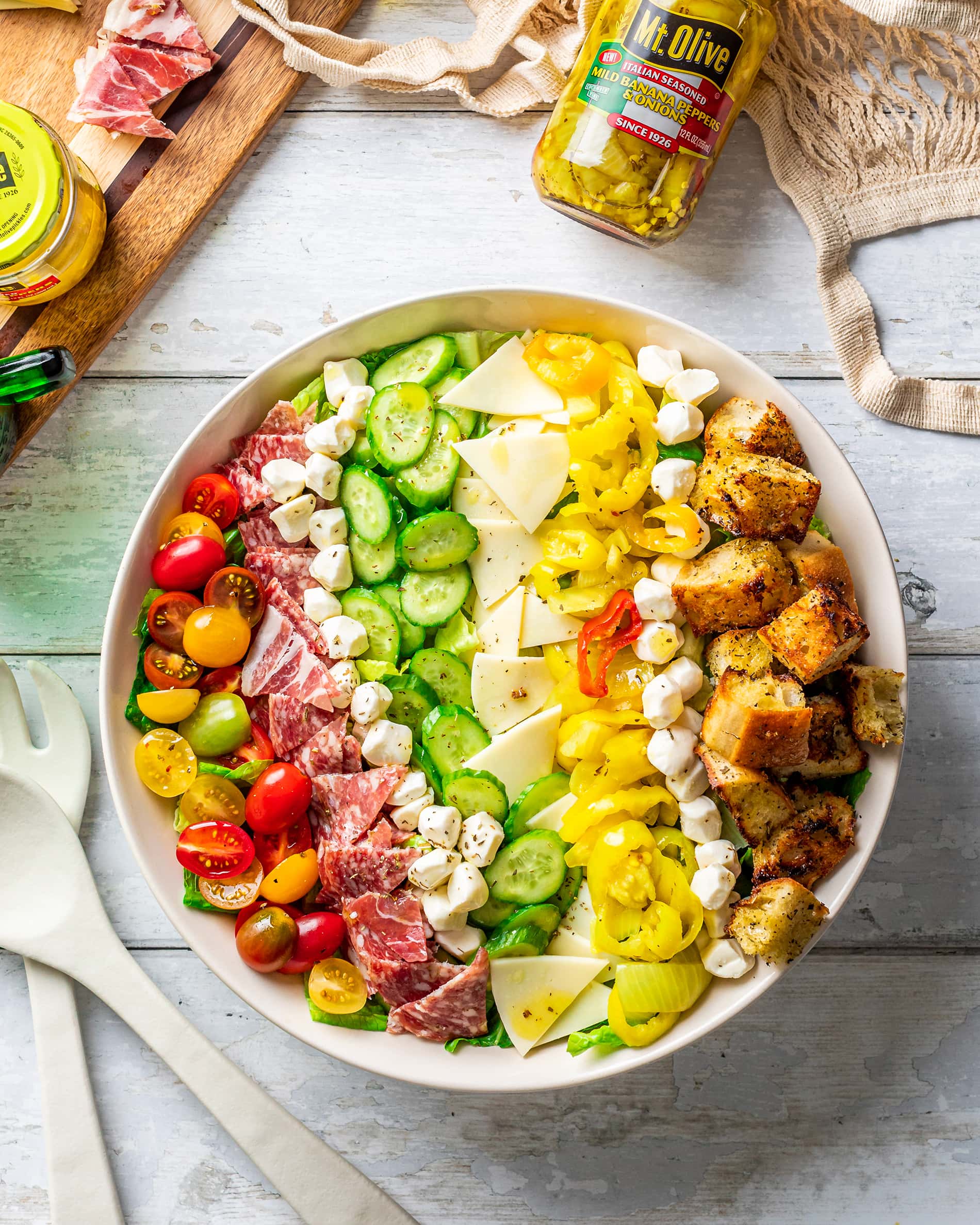 Italian Sub Salad in white bow with white background. Jar of Italian Seasoned peppers at the top. Salad shows tomatoes, meat, cucumbers, peppers and croutons