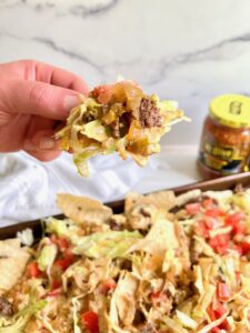 Hand holding tortilla chip with ground beef, cheese, lettuce and pickle salsa - plate of nachos below