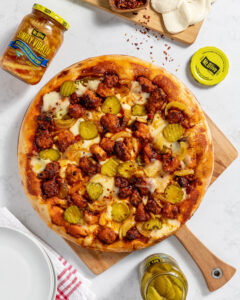 photo is showing a white countertop with a wooden pizza tray. pizza on top is topped with onions pickles and fried chicken. jar of pickles on bottom and jar of pickled onions on top left.