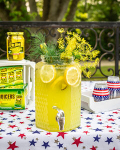 large glass beverage dispenser filled with lemonade and garnished with dill and pickles. 4th of july decor on table. Cases of Pickle Juicers