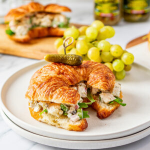 white plate with a chicken salad croissant sandwich topped with a petite dill. green grapes in background. another sandwich in background and two jars of Mt. Olive products in background
