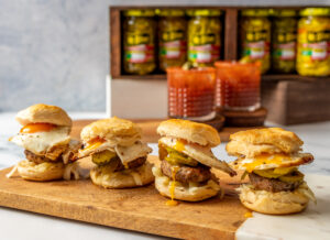 wood board with four spicy breakfast biscuits. Have egg cheese sausage and jalapenos on them. jars in the background and cups with red blood mary