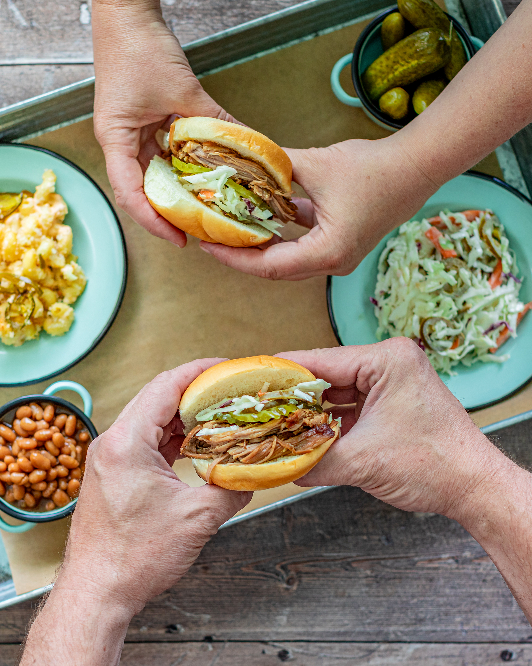 two hands holding pulled pork sandwiches, side dish of cole slaw, beans and mac and cheese