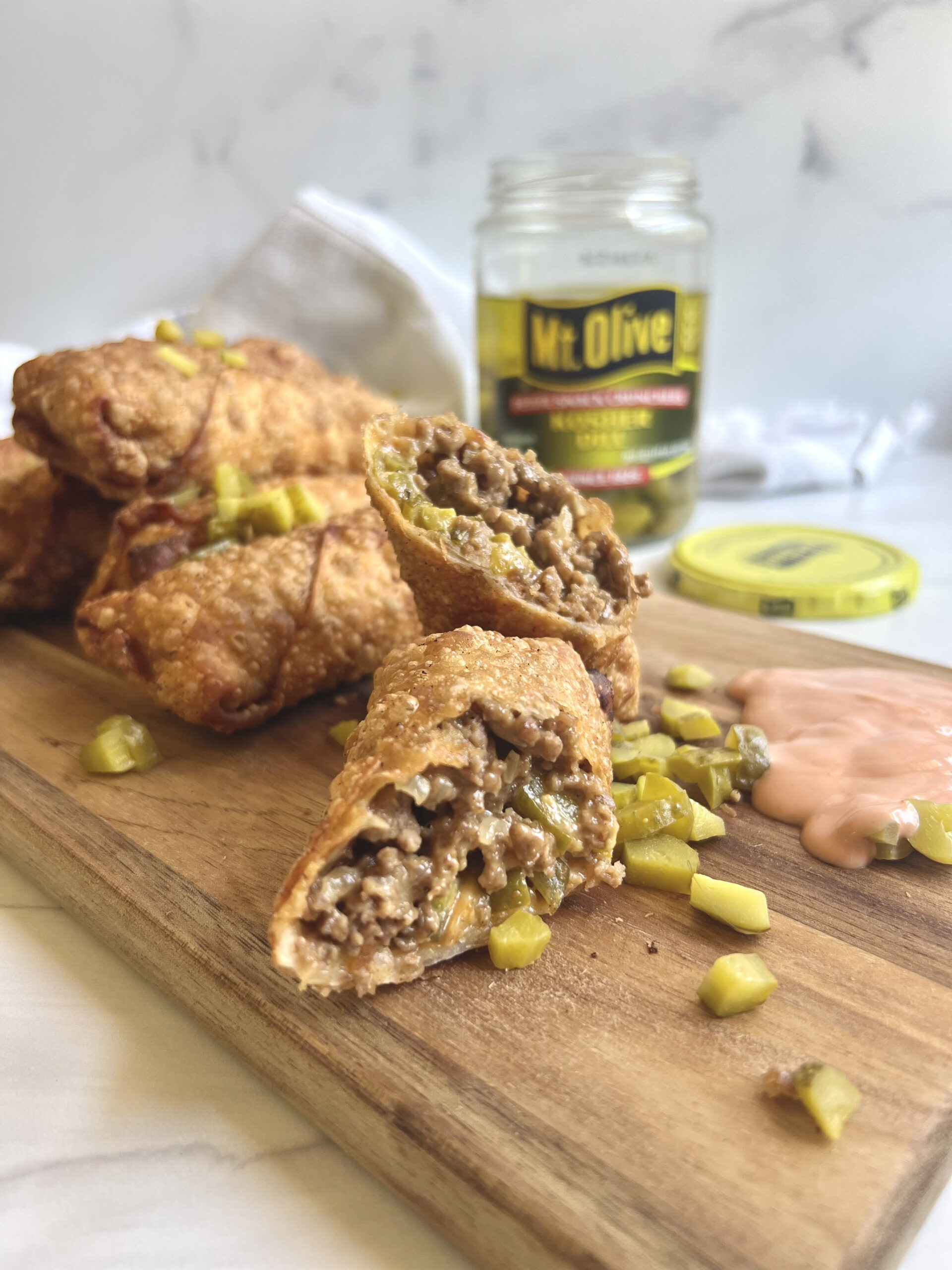 Cheeseburger Egg Rolls on a wooden cutting board with pickles and a jar of pickles in back