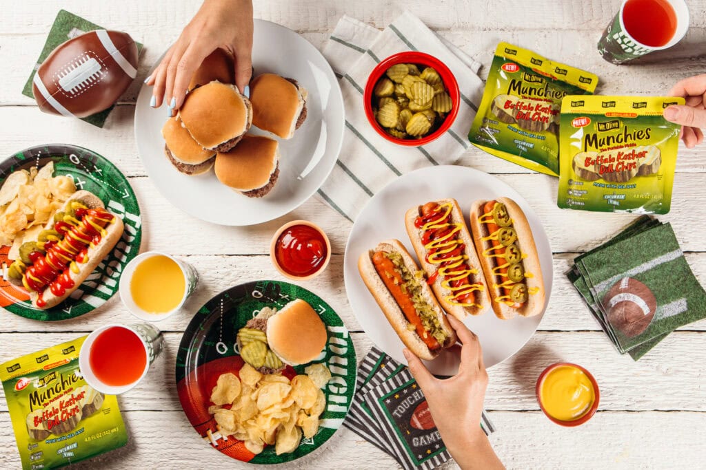 white wood table with plates of burgers and hot dogs showing Buffalo Kosher Dill Chips Munchies Pickle Pouches and ketchup and mustard dishes