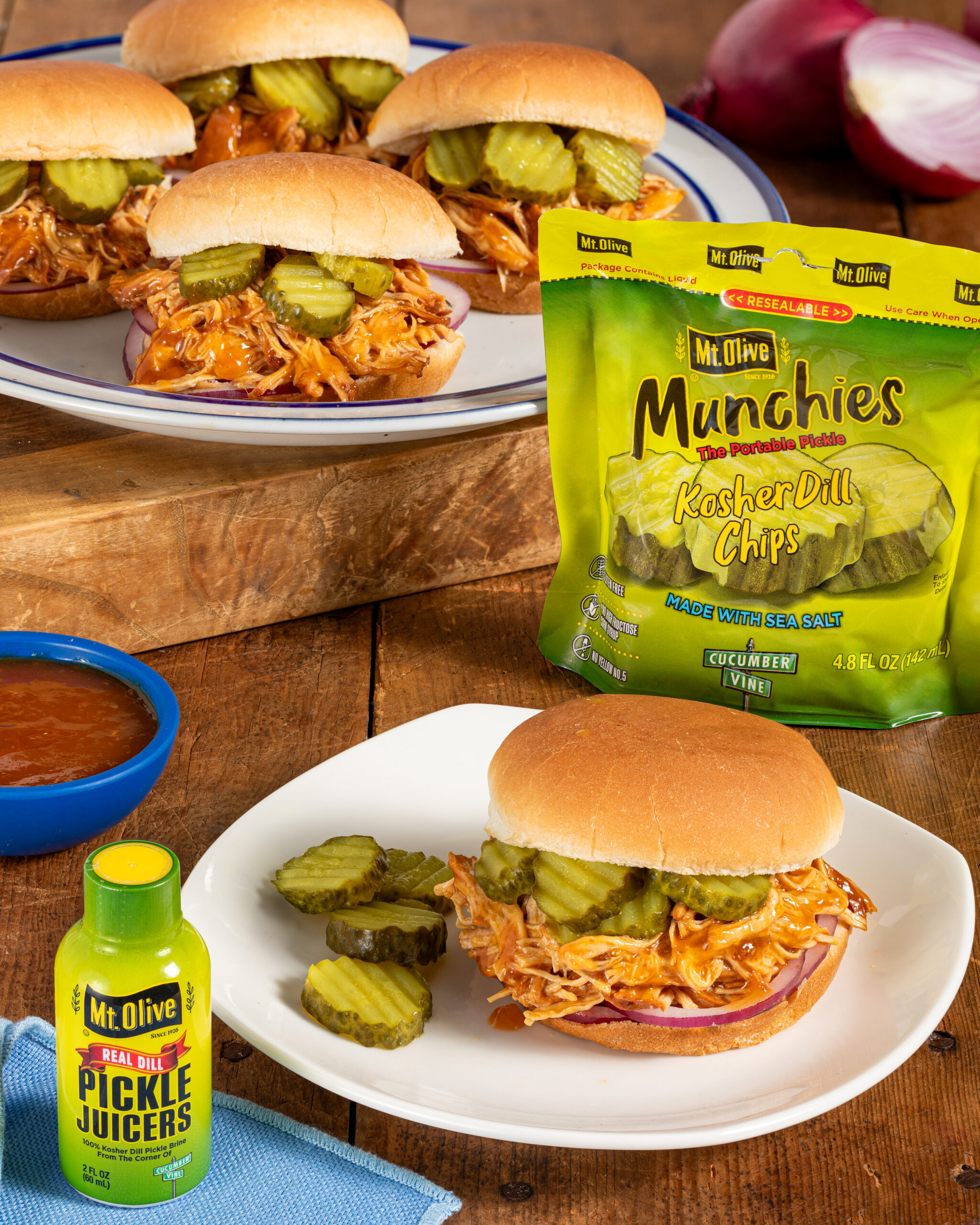 wood table. background shows multiple pulled chicken sliders on buns with pickles. Pouch of portable Munchies Pickles Kosher Dill Chips. White plate shows close up of pulled chicken sandwich topped with pickles. blue dish of bbq sauce. Pickle Juicer bottle in front.