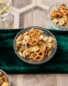 wood table with hunter green tablecloth. glass bowl of seasoned chex mix and pretzels