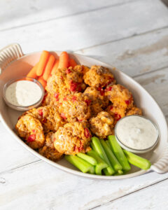 white table with large white platter dish. dish contains celery and carrots as garnish and two small dipping bowls of ranch dressing. middle of bowl contains sausage balls made of roasted red peppers and pimento cheese.