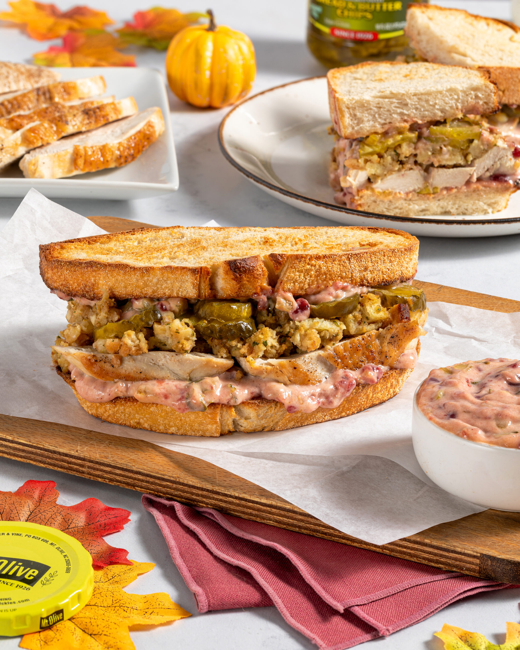 photo shows a large turkey sandwich stacked with leftover thanksgiving turkey meat and stuffing and cranberry pickle mayo with pumpkin and leaf as decor