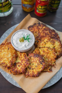 plate of potato pancake latkes on a silver plate with parchment paper lining the plate. in the middle is a dish with pickle sour cream and dill. background shows Mt. Olive jard on wood table