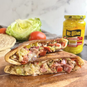 wooden cutting board with an italian stuffed pita. Inside the pita is salami, deli meats, cheese, banana peppers all chopped up and behind is a head of lettuce, a tomato and a jar of Mt. Olive Italian Banana Peppers