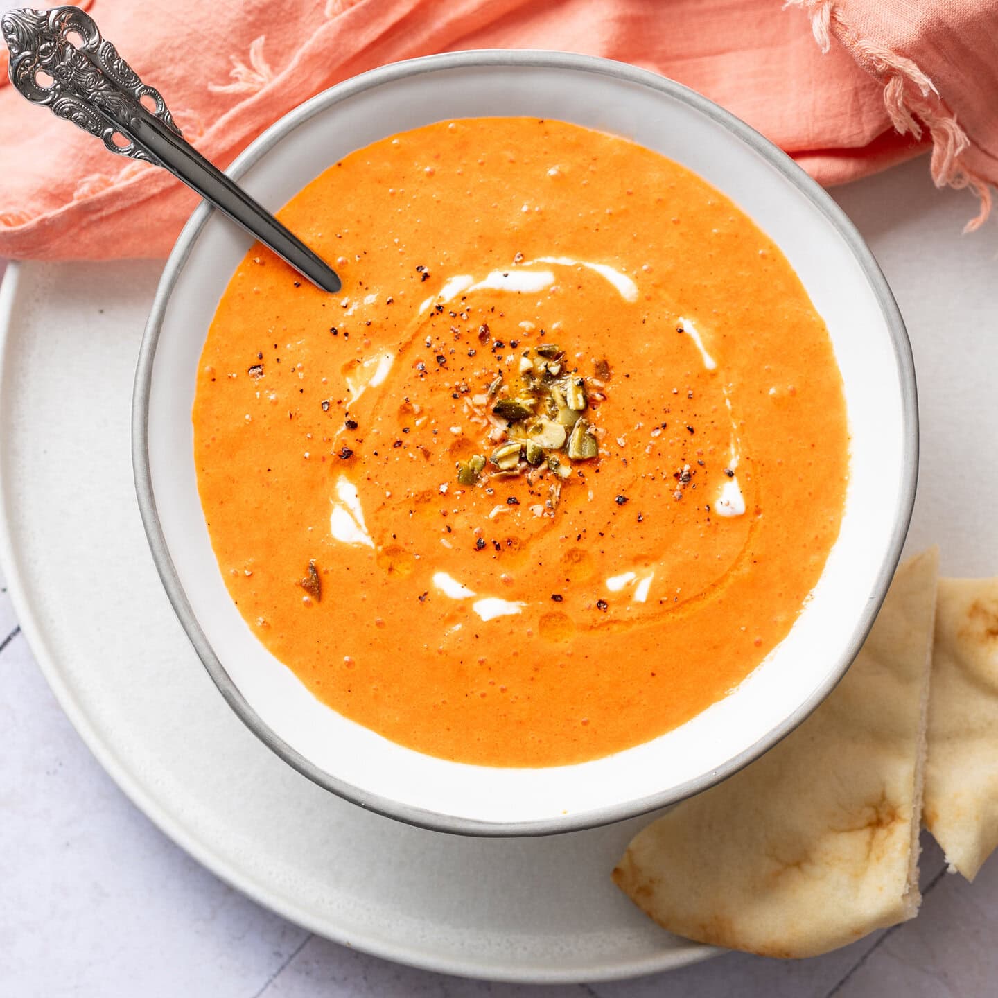white plate with a white bowl - overhead view of bowl of orange soup that is roasted red pepper soup topped with pumpkin seeds