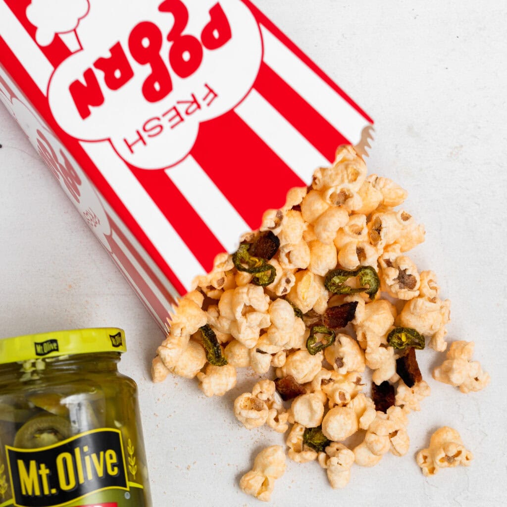 red and white striped popcorn box with popcorn spilling out onto white countertop. popcorn has jalapenos and bacon in it. jar of jalapenos beside it