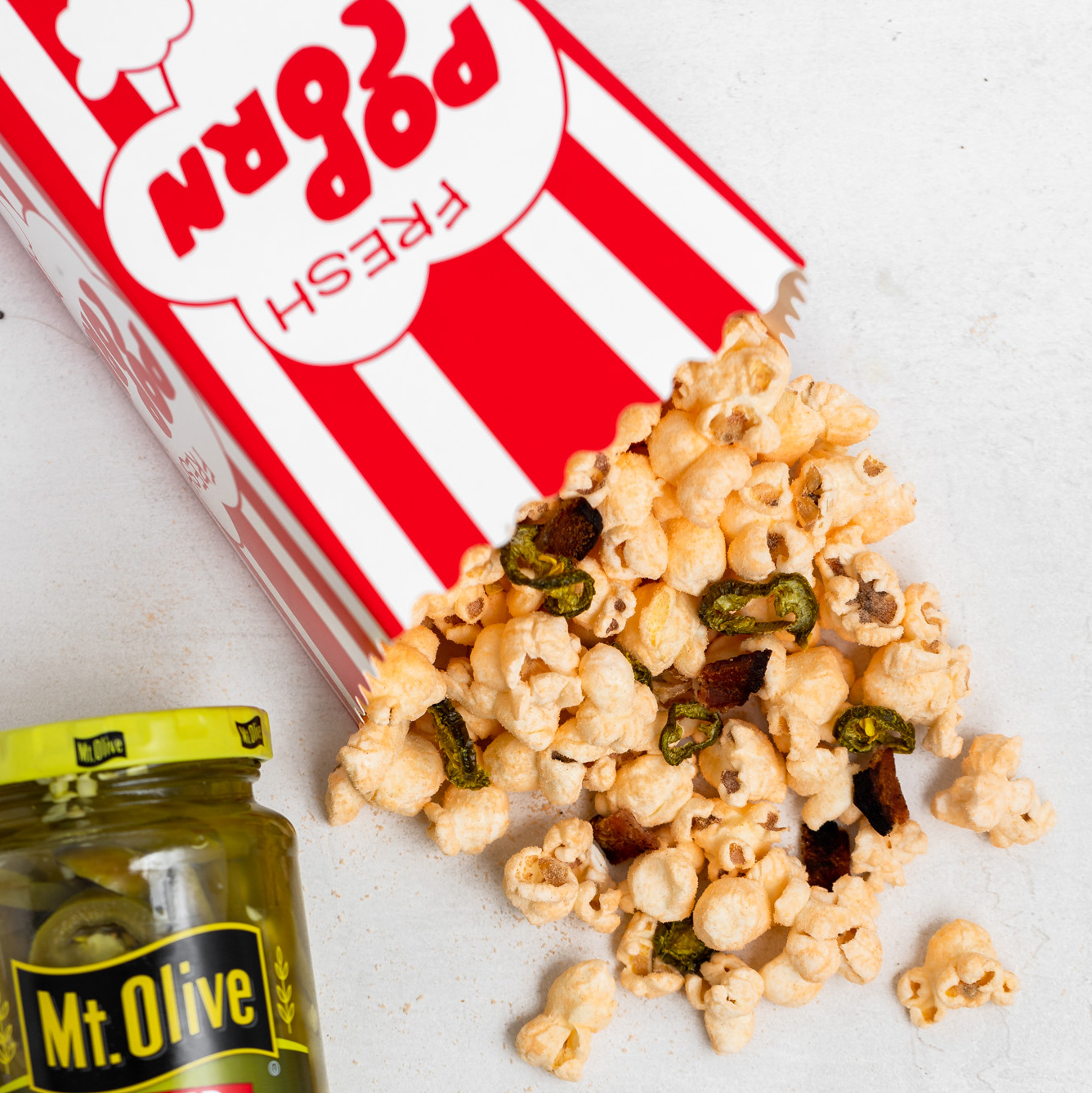 red and white striped popcorn box with popcorn spilling out onto white countertop. popcorn has jalapenos and bacon in it. jar of jalapenos beside it