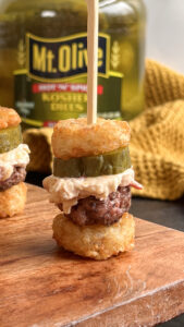 brown wooden butcher block, on top sits a Pimento Cheese Burger Bite, which is made out of tater tots as mini "buns" with a small burger patty, a pickle chip and pimento cheese on top. In the background is a jar of Mt. Olive Pickles