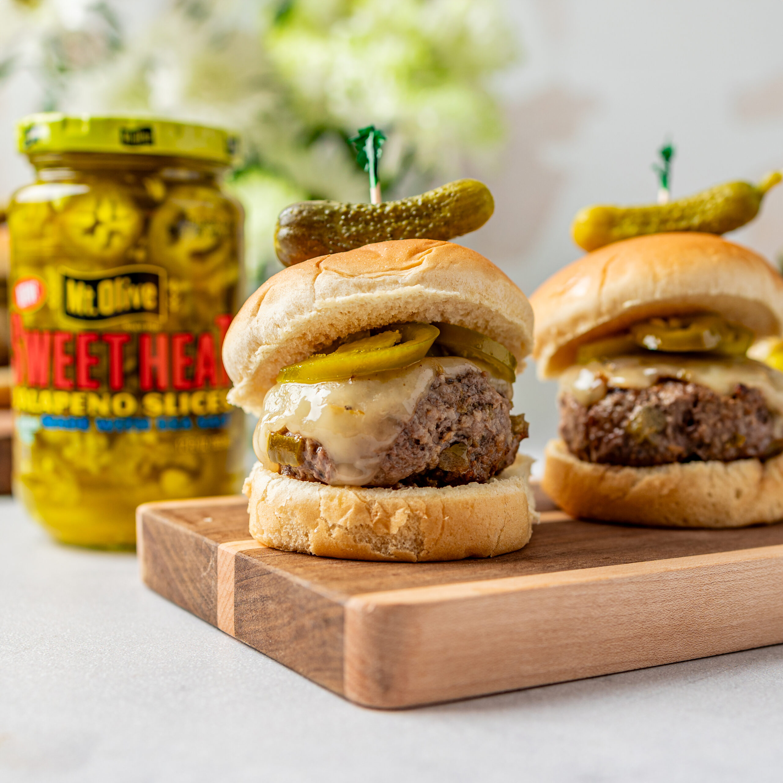 Two sliders (mini burgers) on a wood butcher block with melted cheese and jalapenos, topped with a baby dill pickle. Jar of sweet heat jalapenos behind in background