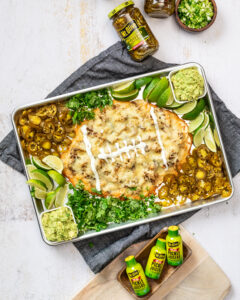 metal pan surrounded by jar of jalapenos and three Mt. Olive Pickle Juicers 2 oz bottles. Pan contains nachos in the shape of a football surrounded by jalapenos and guacamole
