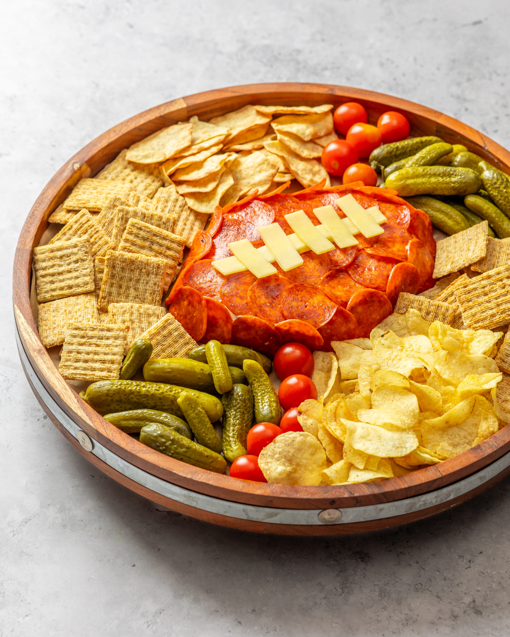 cheeseball covered in pepperoni and cheese to look like a football surrounded by pickles and crackers and chips on a round platter dish