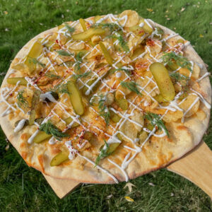 a wood block with grass underneath. has a pizza on top. thin crust topped with pickles and dill