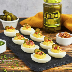 wood table with black tray with 8 deviled eggs topped with bacon bits and pickle slices. jar of pickles in background