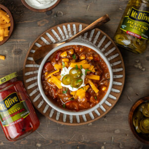 brown table. bowl of chili con carne with a dollop of sour cream and topped with jalapeno slices. jars of roasted red peppers and jalapeno slices to the sides of bowl.