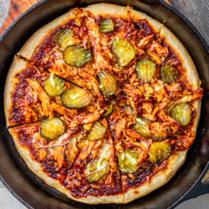 close up of pizza in black cast iron pan. pizza has shredded chicken, barbeque sauce, cheese and pickles on top.